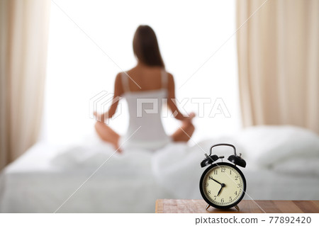 Alarm clock standing on bedside table has already rung early morning to wake up. Woman do yoga in bed in background. Early awakening, healthy lifestyle contemplation concept. Alarm clock standing on bedside table has already rung early morning to wake up. Woman do yoga in bed in background. Early awakening, healthy lifestyle contemplation concept. 77892420
