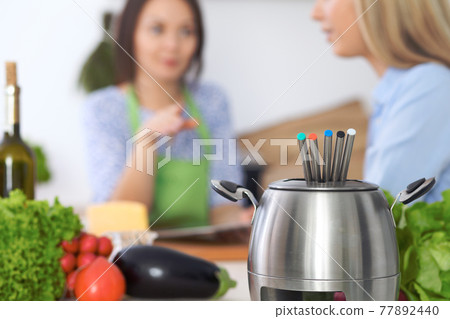 Fondue in a pot at the background of friends cooking together, close-up. Kitchen interiors and cookware Fondue in a pot at the background of friends cooking together, close-up. Kitchen interiors and cookware 77892440