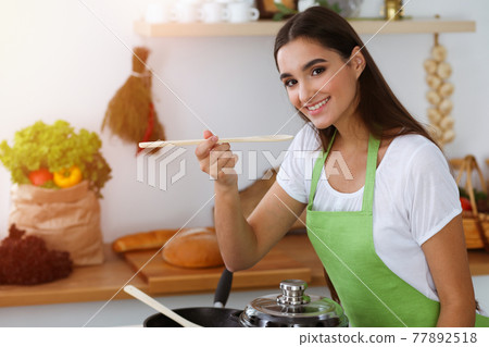 An attractive young dark-haired woman tasting ready hot meal with a wooden spoon while standing and smiling in sunny kitchen. Cooking and householding concepts 77892518