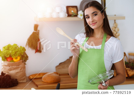 An attractive young dark-haired woman tasting ready hot meal with a wooden spoon while standing and smiling in sunny kitchen. Cooking and householding concepts 77892519