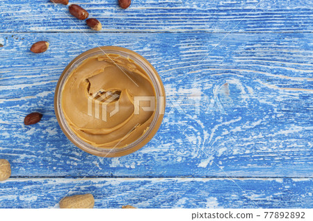 Peanut butter in an open jar and peanuts in the skin are scattered on the blue table Peanut butter in an open jar and peanuts in the skin are scattered on the blue table 77892892