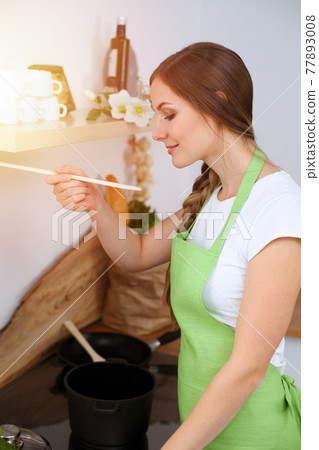An attractive young woman preparing soup by new keto recipe while standing and smiling in sunny kitchen. Cooking and householding concepts 77893008