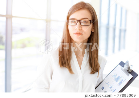 Beautiful female specialist with clipboard standing in sunny office and smiling charmingly. Business people concept 77893009