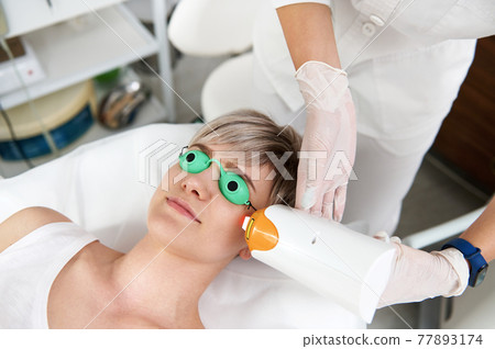 Beautician doing laser rejuvenation in a beauty salon. Elos epilation hair removal procedure on young woman face. Facial skin care. Hardware ipl cosmetology 77893174
