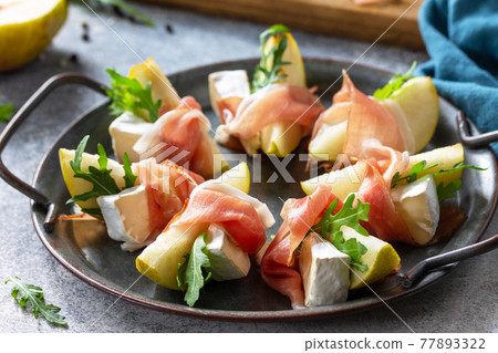 Summer snacks close-up. Pear appetizer with jamon, arugula and brie cheese on a light stone table. 77893322