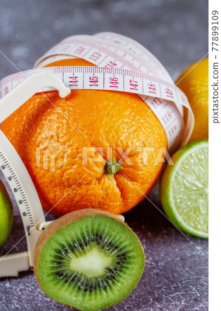 An orange wrapped with a measuring tape and a caliper surrounded by fresh fruits 77899109