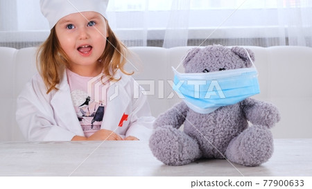 little girl plays a doctor, makes an injection. Happy child little doctor examines a teddy bear 77900633