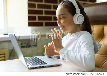 Photo of a pupil girl in white headphones in front of a laptop monitor talking with a teacher during an online lesson. 77906571