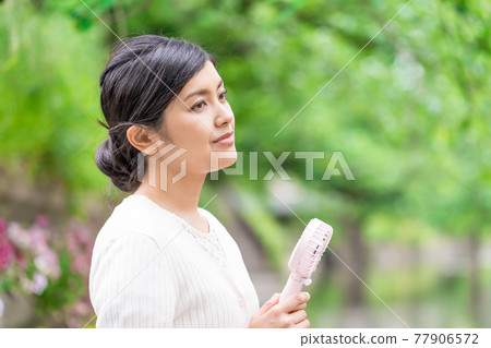 一位女商人在新鮮的綠色植物中拿著一把方便的風扇納涼 一位女商人在新鮮的綠色植物中拿著一把方便的風扇納涼 77906572