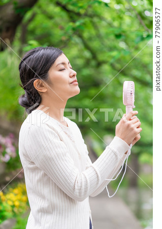 一位女商人在新鮮的綠色植物中拿著一把方便的風扇納涼 一位女商人在新鮮的綠色植物中拿著一把方便的風扇納涼 77906577