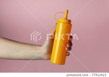 Female hand hold yellow sauce bottle on pink background Female hand hold yellow sauce bottle on pink background 77912623