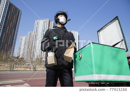 Asian food delivery male courier with mask checking location with smartphone on motorcycle, scooter Asian food delivery male courier with mask checking location with smartphone on motorcycle, scooter 77913288
