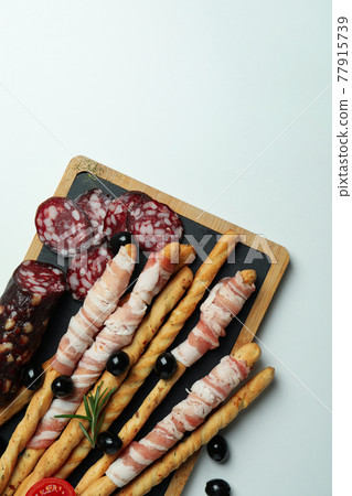 Board with grissini sticks with bacon and snacks on white background Board with grissini sticks with bacon and snacks on white background 77915739