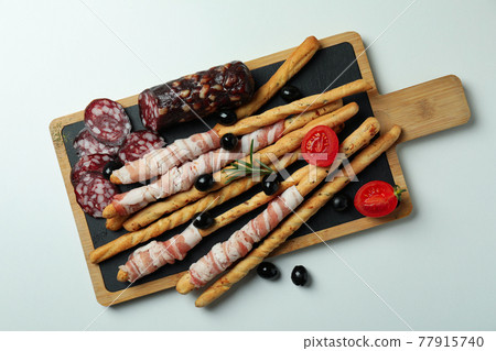 Board with grissini sticks with bacon and snacks on white background Board with grissini sticks with bacon and snacks on white background 77915740