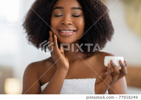 Young African American woman applying hydrating cream on her face, smiling with closed eyes, indoors Young African American woman applying hydrating cream on her face, smiling with closed eyes, indoors 77918249