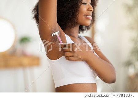 Positive black woman shaving her armpit with razor, removing unwanted body hair at home, selective focus 77918373