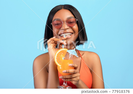 Portrait of cheerful black woman drinking refreshing cool fruit cocktail, having beach party on blue studio background 77918376