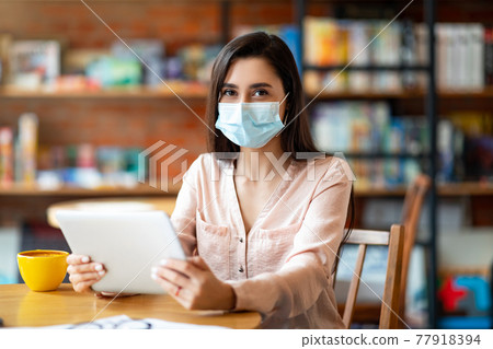 Latin woman in protective mask sitting in coffee shop and using digital tablet, looking at camera Latin woman in protective mask sitting in coffee shop and using digital tablet, looking at camera 77918394
