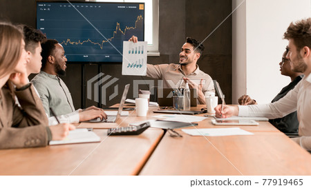 Employee showing paper with graph diagram, sitting at desk 77919465