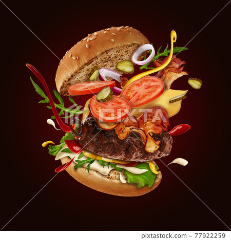 Tasty maxi burger with flying ingredients against brown background. Ham, beef cutlet, cheese, sauces, vegetables and greens. Close up, copy space 77922259