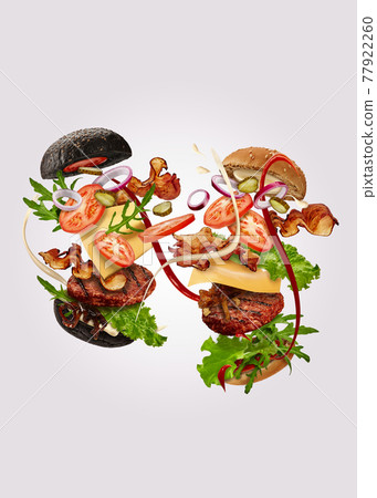 Two burgers, black and classic, with flying ingredients against gray background. Ham, beef cutlet, cheese, sauces, vegetables and greens. Close up Two burgers, black and classic, with flying ingredients against gray background. Ham, beef cutlet, cheese, sauces, vegetables and greens. Close up 77922260