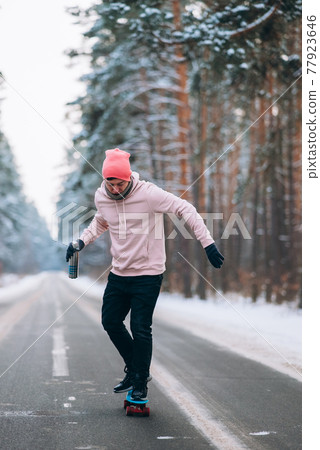 Skateboarder standing on the road in the middle of the forest, surrounded by snow Skateboarder standing on the road in the middle of the forest, surrounded by snow 77923646