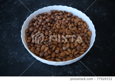 A wooden bowl with mixed nuts on top of a black concrete table. Healthy food and snacks. Nuts, pistachios, almonds, hazelnuts. 77926552