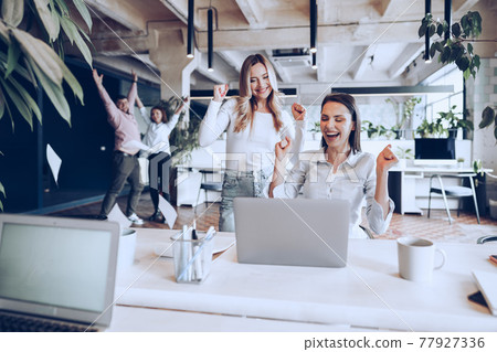 Two young happy businesswomen celebrating project success in office Two young happy businesswomen celebrating project success in office 77927336