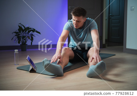Stretching exercises at home, a young guy watches a video on a tablet and repeats the exercises 77928612