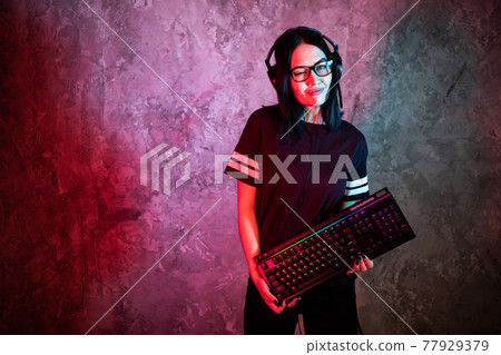 Female esports gamer posing with a gaming gear in neon light. Streamer girl standing with a gaming keyboard. Female esports gamer posing with a gaming gear in neon light. Streamer girl standing with a gaming keyboard. 77929379