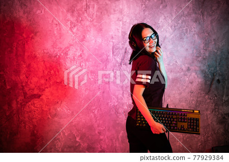 Female esports gamer posing with a gaming gear in neon light. Streamer girl standing with a gaming keyboard. Female esports gamer posing with a gaming gear in neon light. Streamer girl standing with a gaming keyboard. 77929384