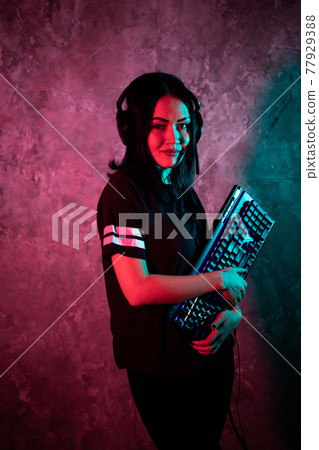 Female esports gamer posing with a gaming gear in neon light. Streamer girl standing with a gaming keyboard. Female esports gamer posing with a gaming gear in neon light. Streamer girl standing with a gaming keyboard. 77929388