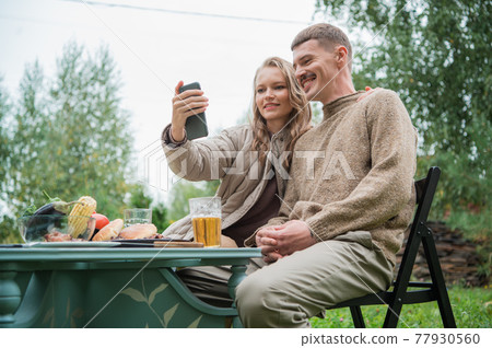 A young couple makes a very funny and emotional selfie, fooling around and making faces in front of the smartphone camera. They made an outdoor dinner with a grill and beer. A young couple makes a very funny and emotional selfie, fooling around and making faces in front of the smartphone camera. They made an outdoor dinner with a grill and beer. 77930560