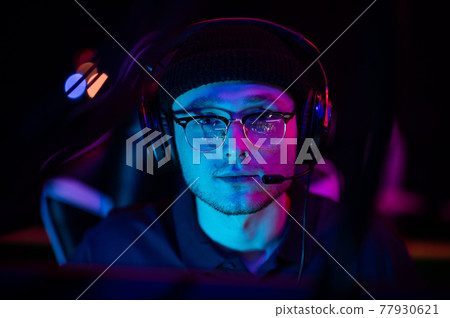 Close-up portrait of a young gamer at the computer. A gaming headset with headphones on the head. Neon light. The concept of esports. 77930621