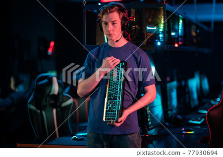 In the computer room, a cool gamer in a headset shows a new wireless keyboard with a backlight, turns it in his hands In the computer room, a cool gamer in a headset shows a new wireless keyboard with a backlight, turns it in his hands 77930694