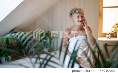 Happy senior woman wrapped in a towel in bathroom at home, resting with tea and smartphone. Happy senior woman wrapped in a towel in bathroom at home, resting with tea and smartphone. 77930909