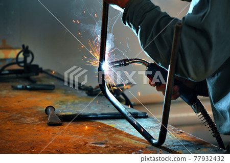 This is a welder working in a welding shop and sparks are flying. This is a welder working in a welding shop and sparks are flying. 77932432