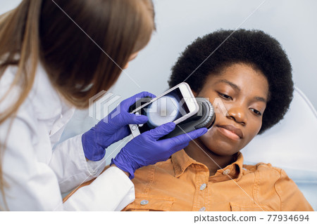 Close up of hands of female doctor holding dermatoscope for study of moles, birthmarks, warts, checking her patient's face. Beautiful afro american woman lying on the couch. Close up of hands of female doctor holding dermatoscope for study of moles, birthmarks, warts, checking her patient's face. Beautiful afro american woman lying on the couch. 77934694
