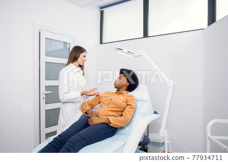 Young African American woman at beauty cosmetology clinic sitting on medical chair, while female doctor wearing white uniform explaining skin treatment procedures. Young African American woman at beauty cosmetology clinic sitting on medical chair, while female doctor wearing white uniform explaining skin treatment procedures. 77934711