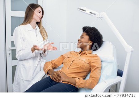 Consultation in cosmetology clinic. Female professional beauty doctor talking with pretty smiling African woman patient and telling good news about skin care and treatment. Skin care and cosmetology Consultation in cosmetology clinic. Female professional beauty doctor talking with pretty smiling African woman patient and telling good news about skin care and treatment. Skin care and cosmetology 77934715