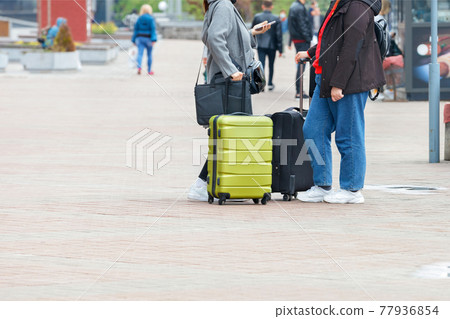 A couple of travelers with suitcases on wheels stand on the sidewalk checking their way on their smartphone. 77936854