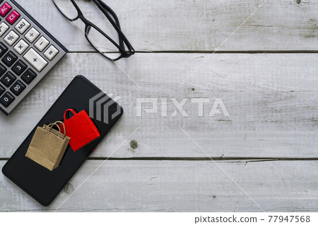 Online shopping concept with shopping paper bags on smartphone, glasses and calculator on white wood table on top view Online shopping concept with shopping paper bags on smartphone, glasses and calculator on white wood table on top view 77947568