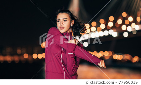 Young fitness woman in headphones stretching her arm at night during a workout Young fitness woman in headphones stretching her arm at night during a workout 77950019