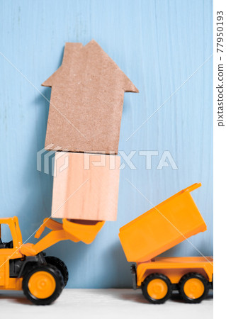 Orange toy excavator loads wooden cube and cardboard house into back of truck. 77950193