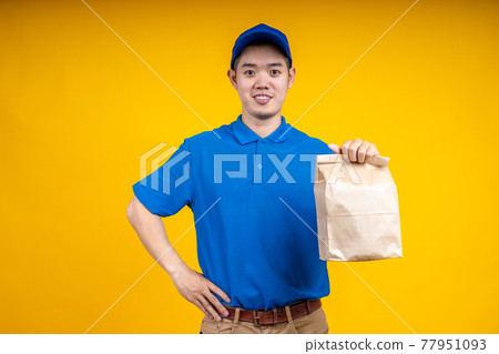 Young Asian handsome delivery man wearing mark holding a food paper bag on right hand over yellow isolate background. Work from home and delivery concept. Young Asian handsome delivery man wearing mark holding a food paper bag on right hand over yellow isolate background. Work from home and delivery concept. 77951093