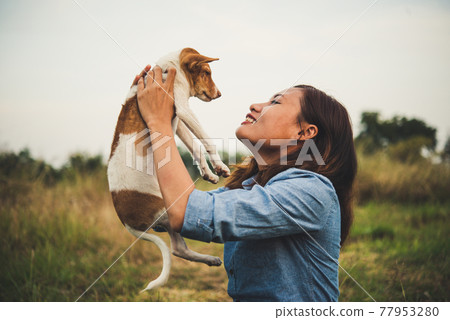 Happy hipster woman smiles and holds a small dog. Little puppy with hipster girls in field sunset background. Vintage tone style. 77953280