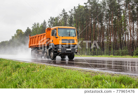 Orange dump truck with 4 axles 40 tonne truck driving on a wet road against the background of a forest, industry. Copy space for text, tonnage Orange dump truck with 4 axles 40 tonne truck driving on a wet road against the background of a forest, industry. Copy space for text, tonnage 77965476