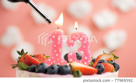 Birthday cake number 13, pink candle on beautiful cake with berries and lighter with fire against background of white clouds and pink sky. Close-up Birthday cake number 13, pink candle on beautiful cake with berries and lighter with fire against background of white clouds and pink sky. Close-up 77969592