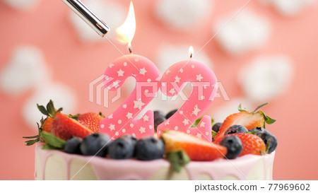 Birthday cake number 22, pink candle on beautiful cake with berries and lighter with fire against background of white clouds and pink sky. Close-up Birthday cake number 22, pink candle on beautiful cake with berries and lighter with fire against background of white clouds and pink sky. Close-up 77969602