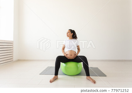 A young pregnant woman doing relaxation exercise using a fitness ball while sitting on a mat. A young pregnant woman doing relaxation exercise using a fitness ball while sitting on a mat. 77971252